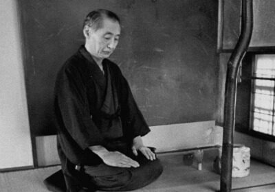 A man in a kimono meditates on the floor in front of a pot.