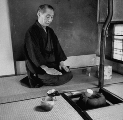 A man in a kimono meditates on the floor in front of a pot.