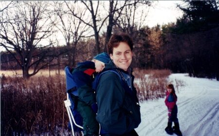 A man venturing on a snowy trail, holding a child in a backpack.