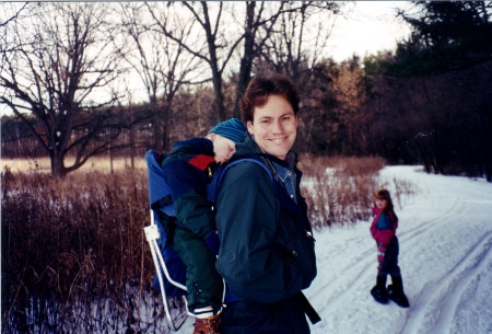 A man venturing on a snowy trail, holding a child in a backpack.