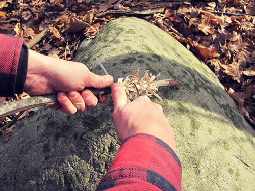 Man using pocket knife to remove bark of stick. 