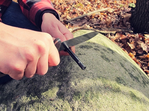 A man using pocket knife for fire-starting ferro-rod.