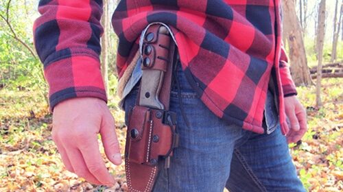 A man wearing a plaid shirt standing in the woods, carefully choosing the perfect survival knife.