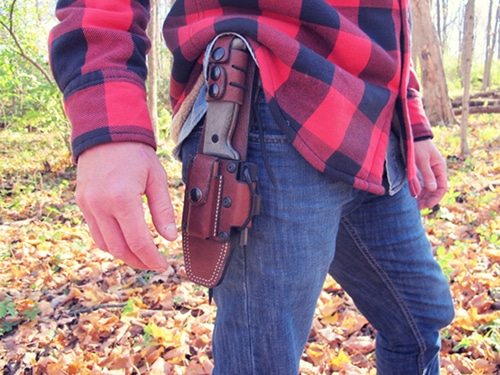 A man wearing a plaid shirt standing in the woods, carefully choosing the perfect survival knife.