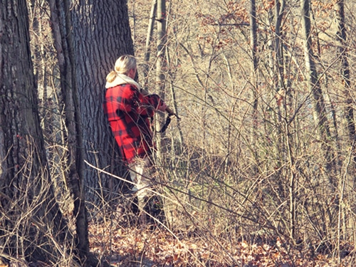 Man jaagt in bos met geweer en draagt rode plaid.