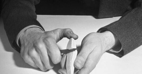 A black and white photo of a man whittling a piece of paper, demonstrating the beginner's guide.