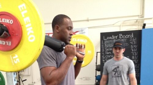Two men standing in a gym, demonstrating squats and barbells. The gym owner provides insight into their job responsibilities and experiences.