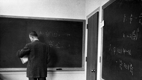 A man in a suit standing in front of a blackboard, demonstrating the formula for success.
