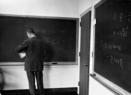 A man in a suit standing in front of a blackboard, demonstrating the formula for success.