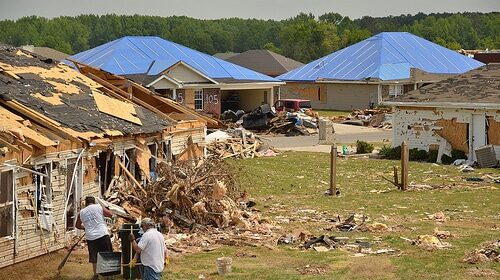Two volunteer men standing in front of a house that has been destroyed by a tornado, providing hands-on disaster relief assistance.