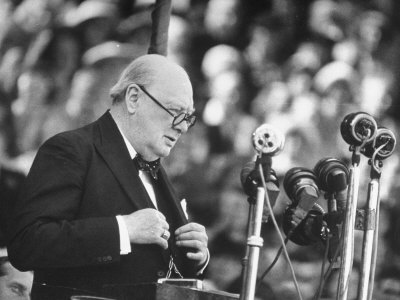 Winston Churchill hält eine Rede mit Brille und Mikrofon.