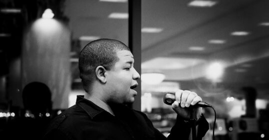 A man, who is also a poet, passionately sings into a microphone in front of a window as he pursues his artistic ambitions.
