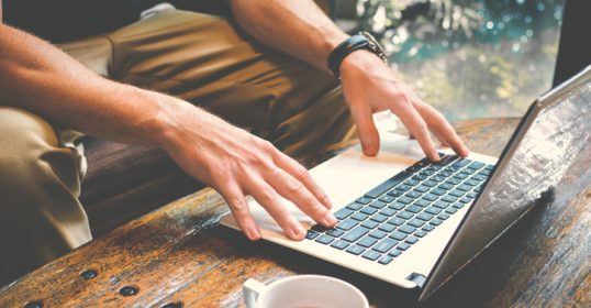 A man writing on a laptop with a cup of tea.