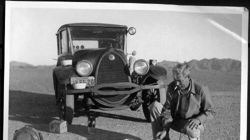 A man taking an impulse road trip, kneeling next to a car in the desert.