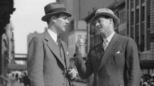 Two men in suits talking on the street, engaged in a conversation focused on crafting good questions and actively listening to each other's responses.