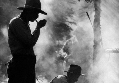 Black and white photograph of two men cooking with cast iron in the woods.