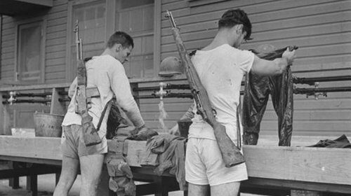 Two men in shorts standing on a bench with rifles.