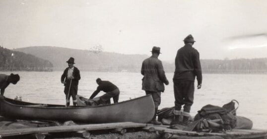 A group of men embarking on a Wilderness Canoe Trip standing on a log next to a canoe, ready to Paddle Away.