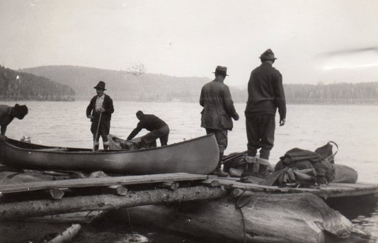 A group of men embarking on a Wilderness Canoe Trip standing on a log next to a canoe, ready to Paddle Away.
