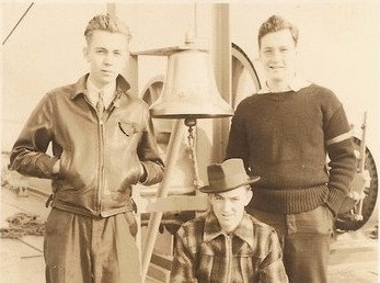 Three young men standing next to a bell on a ship.