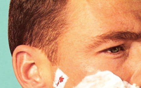 A man is shaving his face with shaving cream and treating any cuts with toilet paper.