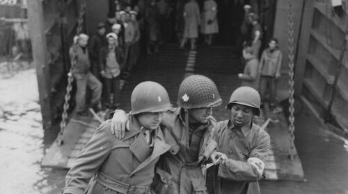 A group of soldiers, led by a combat medic, are bravely walking on a bridge amidst the chaos of battle. Equipped to save lives, the medic remains vigilant for any signs of