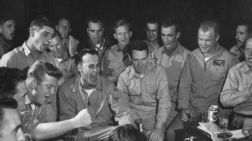 A round of men sitting around a piano, enjoying drinks.