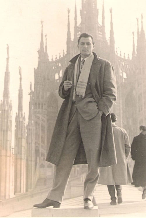A man's guide, dressed in an overcoat, standing in front of a building.