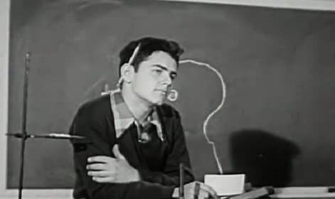 A vintage man sitting at a desk in front of a blackboard, showcasing an instructional film worth watching.