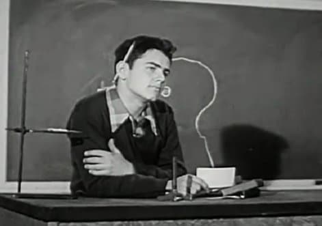 A vintage man sitting at a desk in front of a blackboard, showcasing an instructional film worth watching.