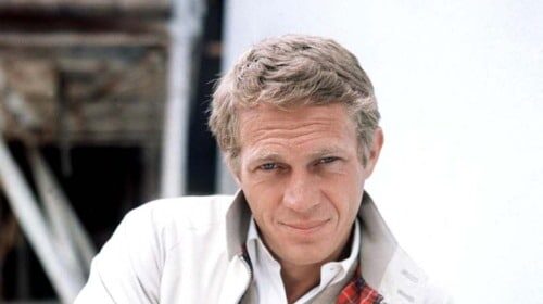 A stylish man in a white jacket leaning against a wall.