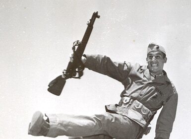 A man undergoing intense training at a boot camp, leaning over a fence with a rifle.