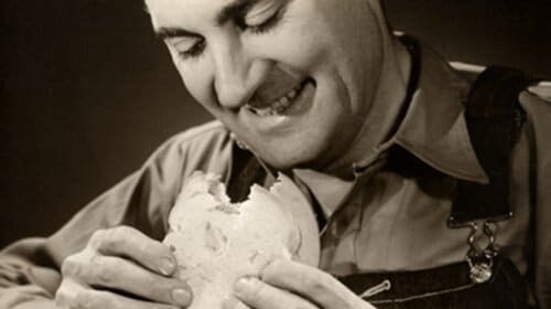 A man in overalls is holding an upgraded humble bologna sandwich.