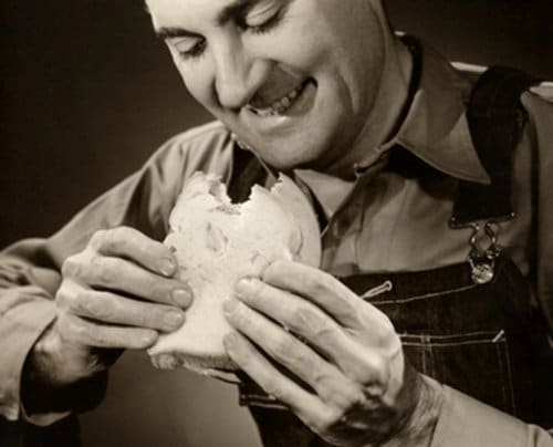A man in overalls is holding an upgraded humble bologna sandwich.