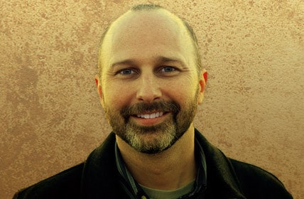 A man smiling in front of a tan wall while recording a podcast.
