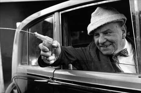 Vintage old man shooting water squirt gun outside car window.