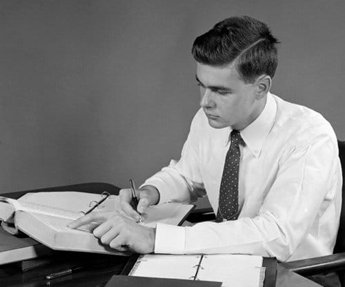 Vintage young businessman at desk looking through textbook taking notes.