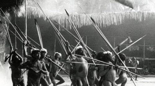A group of Yanomamö tribe members wielding spears, showcasing the origins and male honor of their community in the sandy landscape.