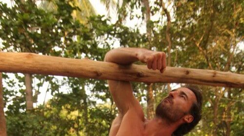 A fit Wild Man performing a handstand on a wooden pole.