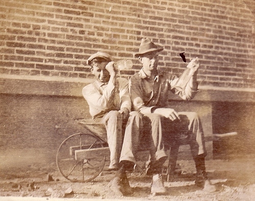 Vintage old time photo two men sitting in wheelbarrow with bottles.