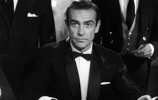 A man in a tuxedo with a bow tie sitting at a black-tie event, surrounded by others in formal attire.