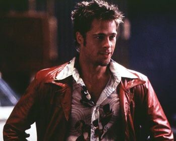 Brad Pitt, wearing a red leather jacket, is standing in front of a car.