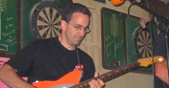 A guitarist playing an orange guitar in front of a dart board.