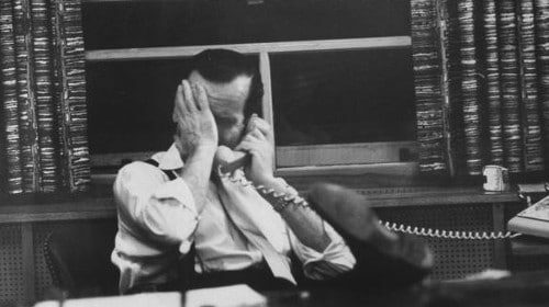 A man sitting at a desk discussing a deal on the phone for a job he doesn't like.