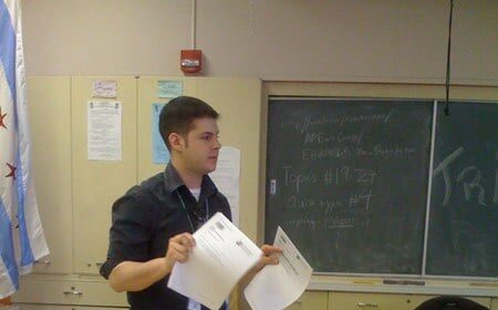 A high school teacher standing in a classroom with papers in front of him.