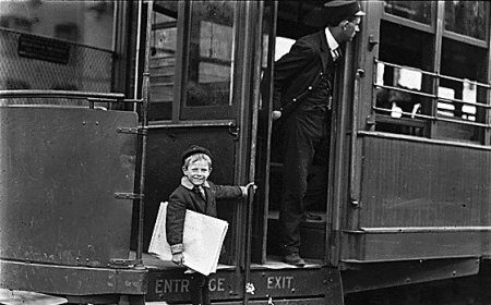 A kid is standing on the door of a trolley car.