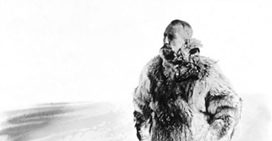 A man in a fur coat standing in the snow on a cold morning.
