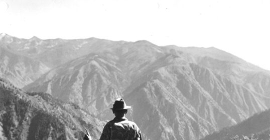 A man standing on top of a mountain with his own rifle for survival reasons.
