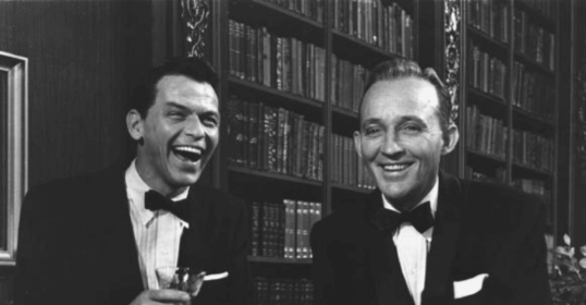 Two men in tuxedos smiling at each other, enjoying gin cocktails.