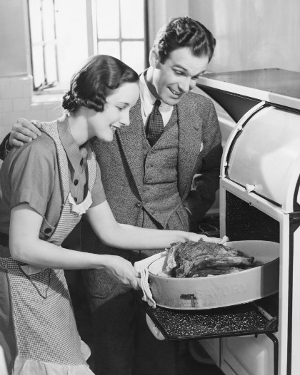 Vintage wife putting roast in oven husband looking on.